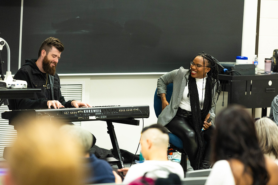 A performing artist smiles as she looks at a keyboard player seated to her right as they perform for college students in a classroom setting.