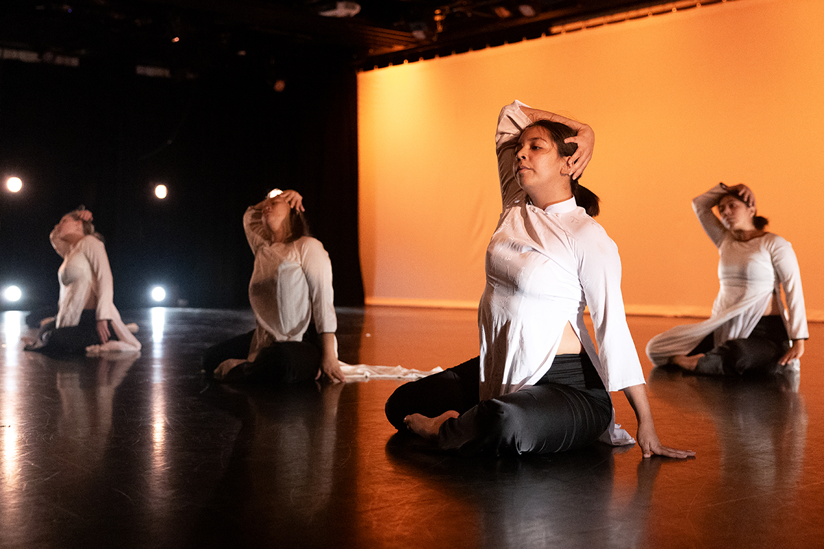 Four dancers sit on the floor during a performance, each placing their right arm on top of their head.