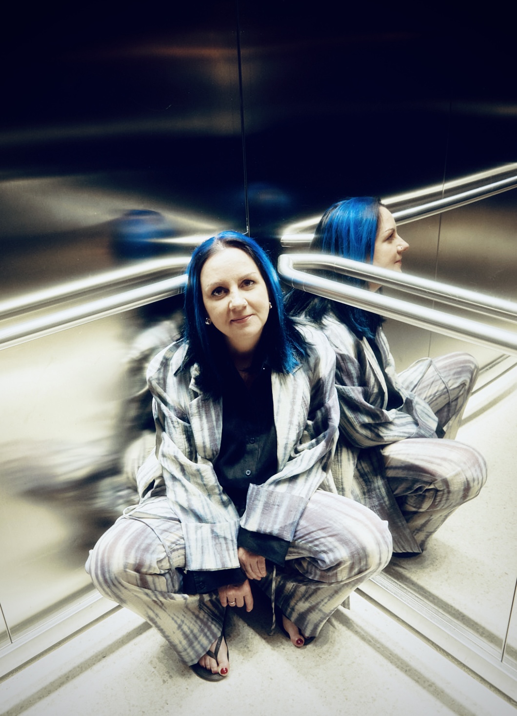 A woman sits crouched in an elevator with her reflection showing in the mirrors behind her.
