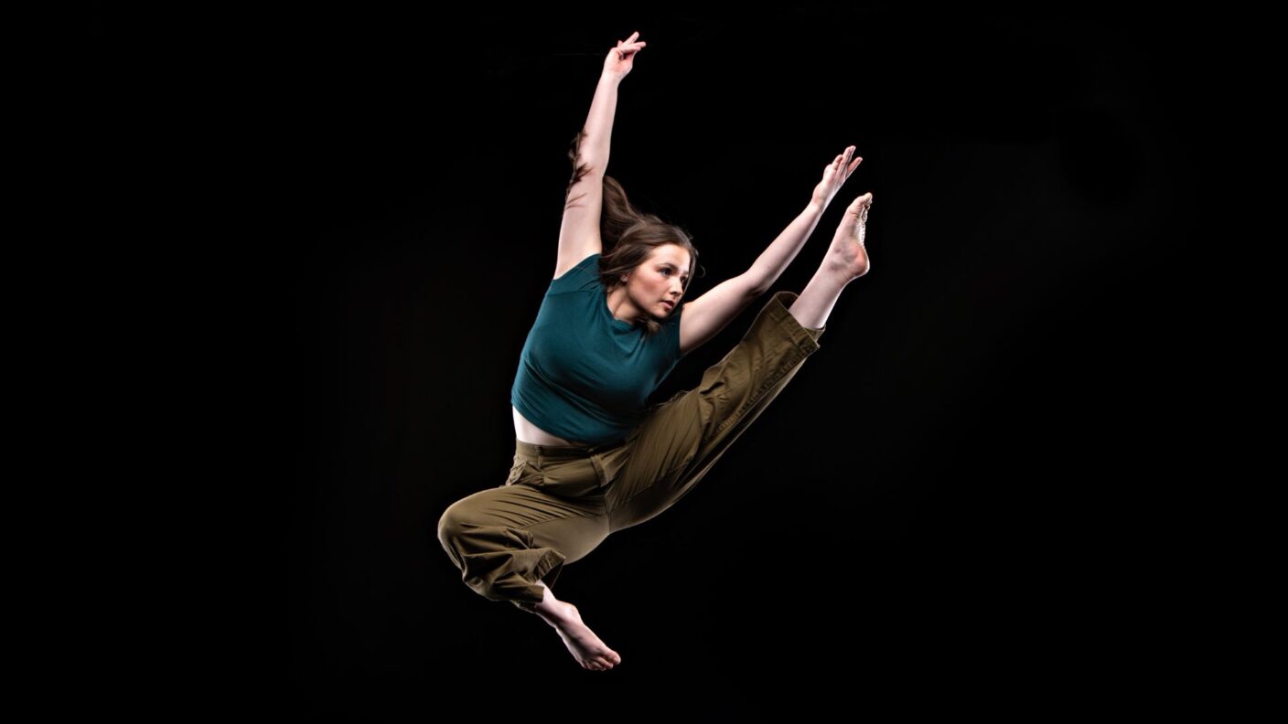 A dancer is shown in in midair, with her arms extended up, and her left leg extended out.