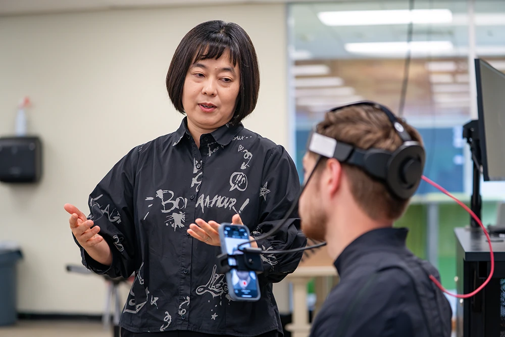 A university professor stands next to a student as he is wearing virtual reality gear that includes a headset with wires, and a cell phone is being held in front of his face that is also attached to the headset.