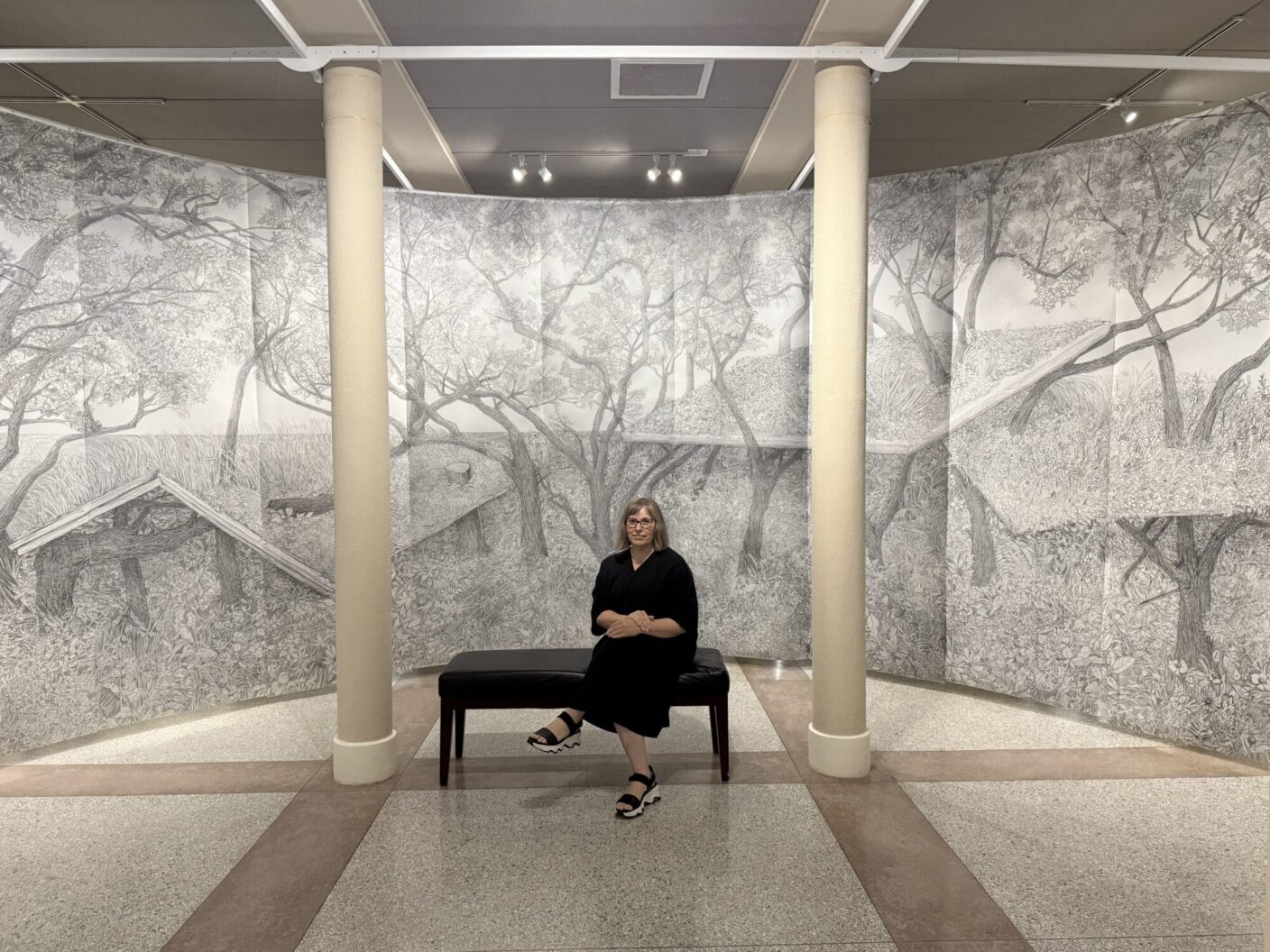 A woman sits on a bench in an art exhibit. Behind her is a large piece of artwork on the wall displaying imsges of trees and brush. She sits between two pillars. 