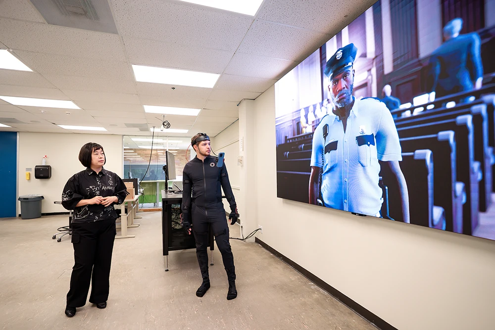 A university professor stands next to a student as he is wearing virtual reality gear that includes a headset with wires. They both stand next to a projector screen on the wall that is showing an image of a police officer. 