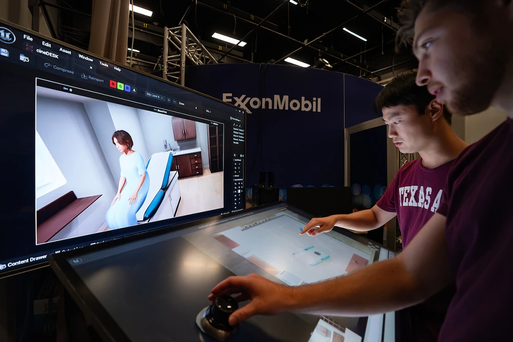 Two university students interact with a touch screen while looking at a computer screen showing a virtual patient in doctors office. Behind them is a large wall that says 'Exxon Mobil.'