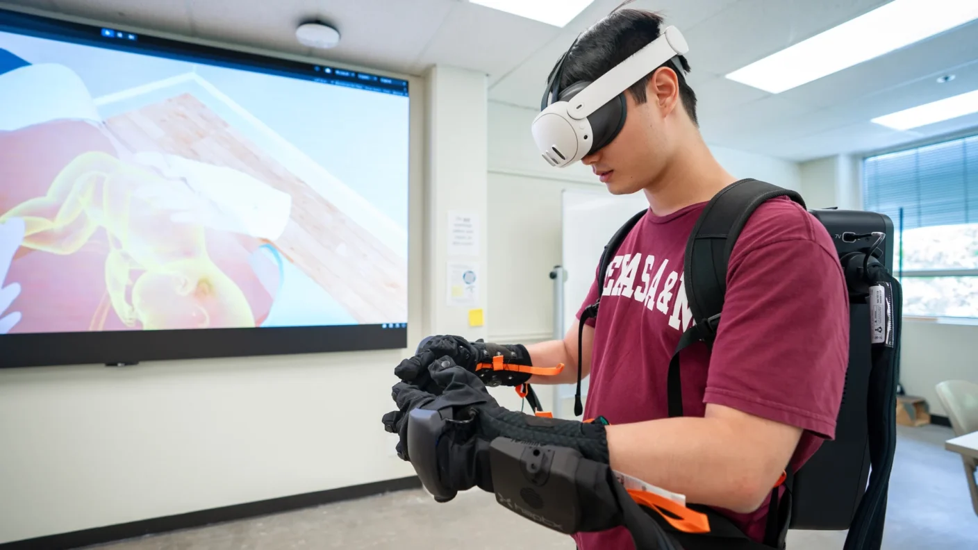 A university student wears virtual reality gear, including a headset, gloves and backpack. he is standing in front of a projector screen that is showing a virtual baby on its mothers stomach. His shirt says 'Texas A&M'
