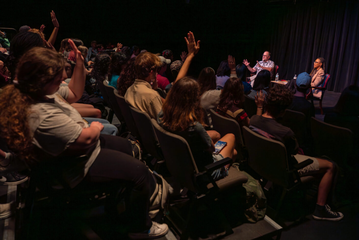 A group of students sit with their hands raised in a Black Box Theatre. 