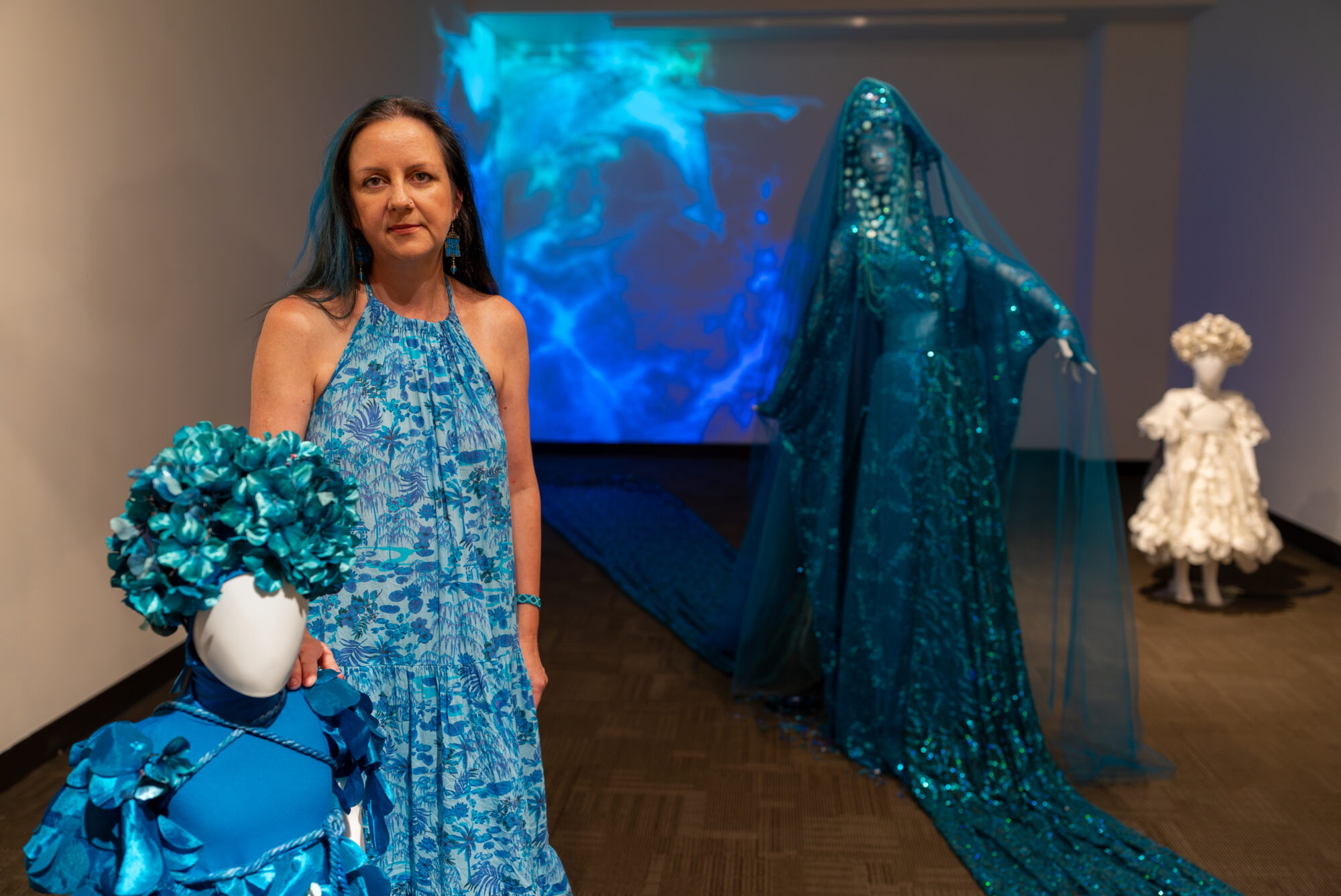 A woman stands next to three mannequins in a unversity art gallery. The mannequins are decorated with detailed costumes.