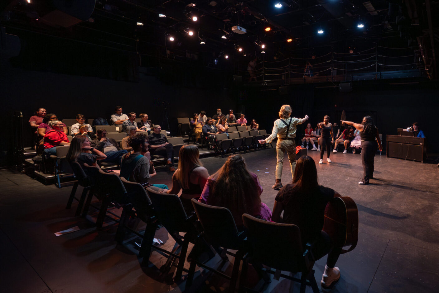 Students in a play rehearse in a black box theatre surrounded by people in theatre seats.
