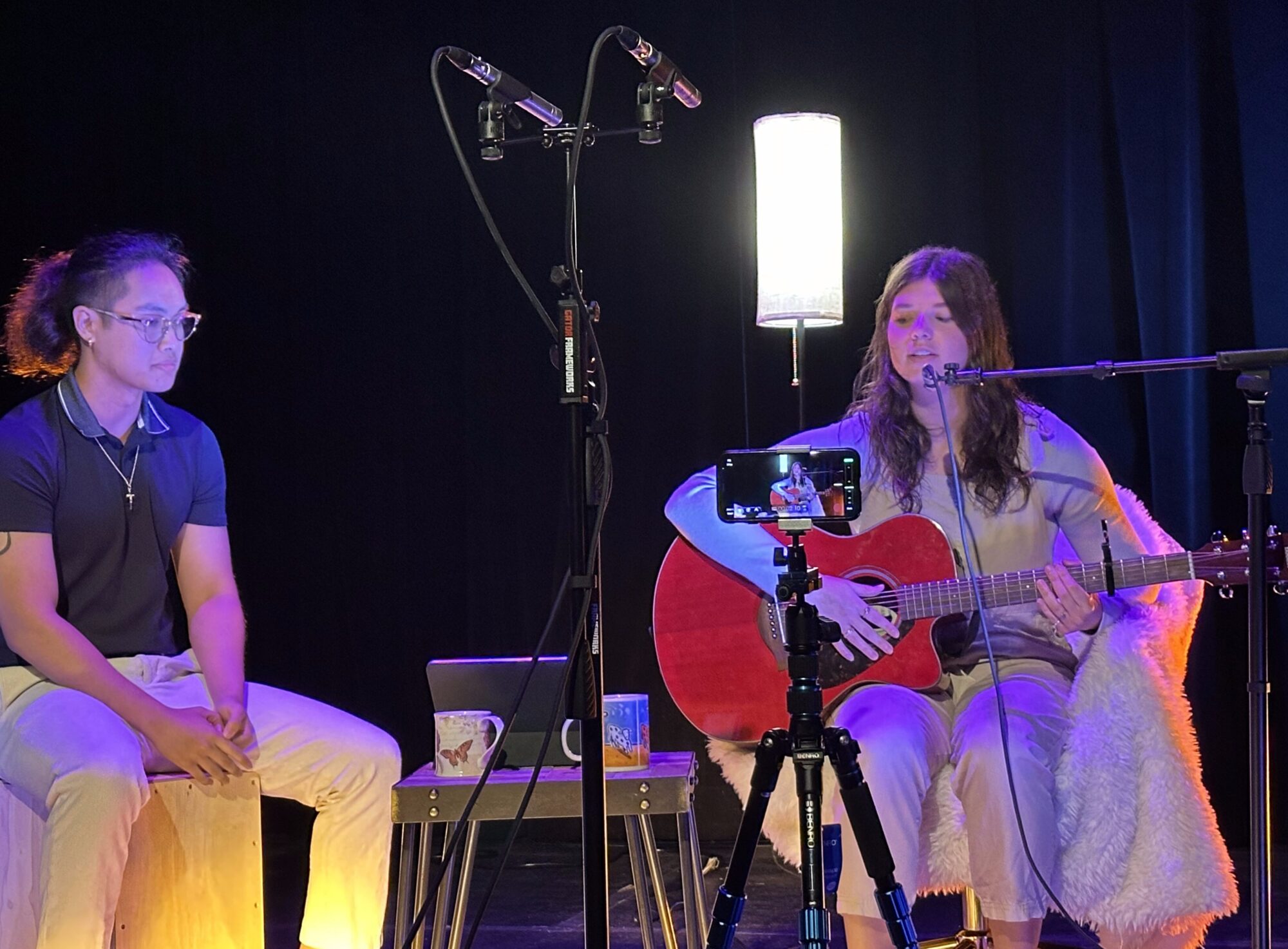 A college student sits and sings into a microphone while playing guitar. At her right is another student accompanying her on percussion.