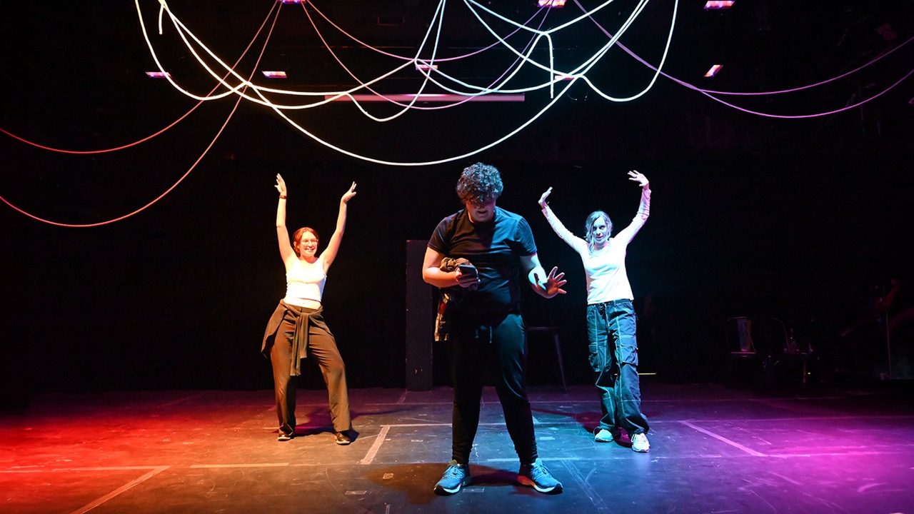 Three performers on a stage with dynamic lighting and an array of hanging cords in the background.