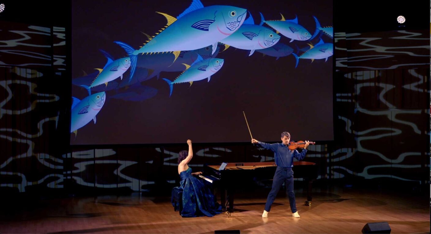 A person playing a violin on stage, with another person partially visible, playing a piano. The background features a large, colorful graphic of stylized fish.