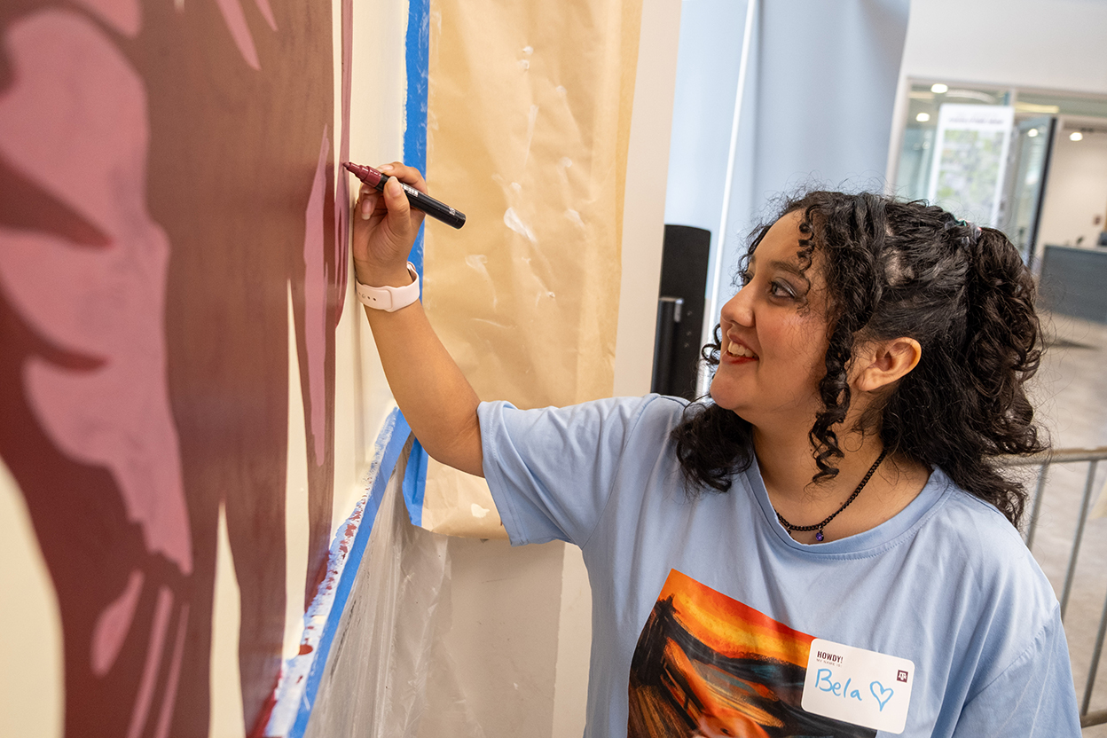 A college student smiles as she adds accents with a paint marker on a wall mural.
