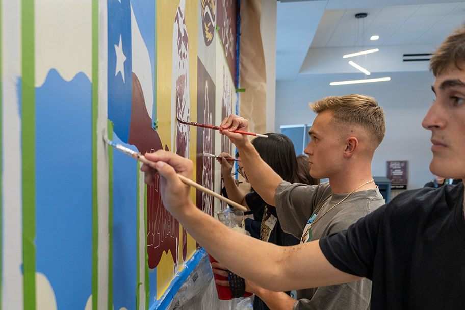 College students paint various scenes on a wall mural.