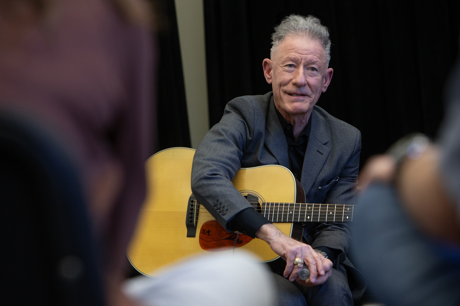 Lyle Lovett sits with a guitar while he speaks to students.