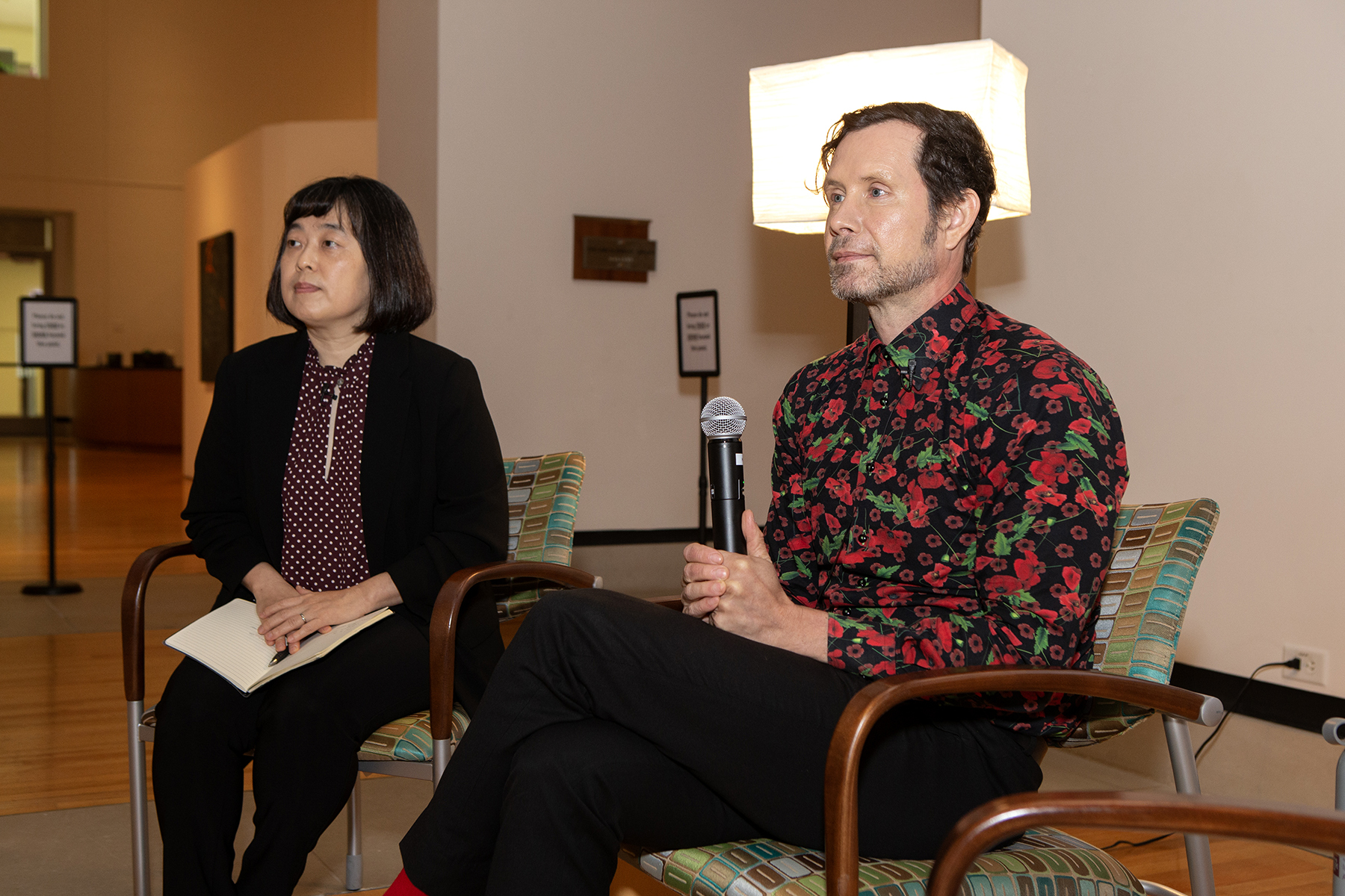 Two people sit in chairs in a university art gallery. The person on the right is holding a microphone, the person on the left is holding a book. Behind them is a lamp.