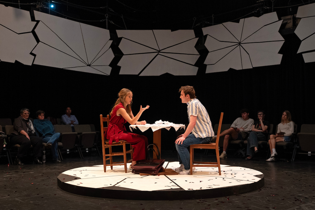 A stage scene with two individuals seated at a round table, engaged in conversation. The table has a white tablecloth with some scattered papers. The background features geometric white panels, and there is a small audience seated in the shadows.