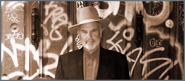 A black and white photo portrait of a man in a cowboy hat, standing in front of a wall filled with graffiti.